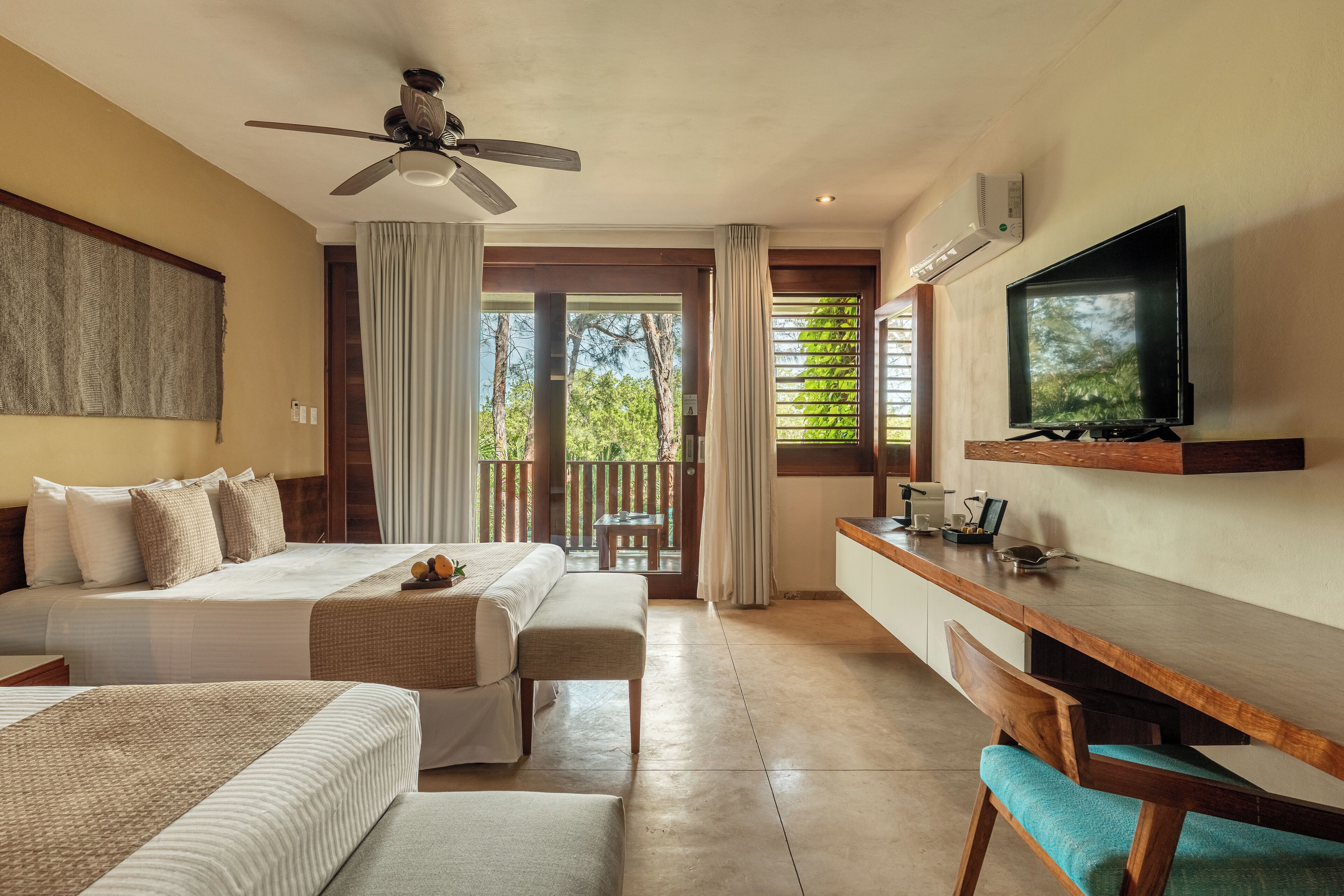 Une chambre d’hôtel avec deux lits, un bureau, une télévision et un balcon donnant sur un paysage arboré.