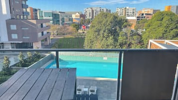 A balcony with a view of a swimming pool, modern buildings, and a grassy area.
