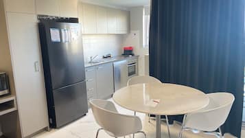 A modern kitchen with a black refrigerator, white cabinets, a sink, and a stove. There is a round table with two chairs and a small shelf with a microwave.