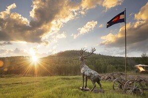 Una estatua de ciervo en un campo cubierto de hierba, con un asta y una bandera, frente a una colina boscosa y un atardecer.