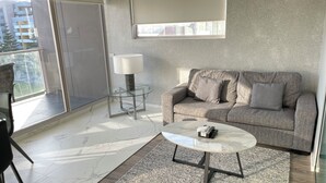 A modern living room with a sofa, a glass coffee table, a lamp, and a view of a balcony. - Premium One Bedroom Apartment (Newcastle)