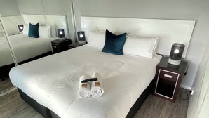 Un lit bien fait, avec du linge de lit blanc, une télécommande et des serviettes posées sur le lit. Il y a deux tables de chevet avec des lampes et une petite armoire.