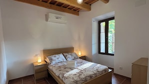 A bedroom with a wooden bed, two bedside tables with lamps, a window with a view of greenery, and a ceiling fan.