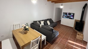 A living room with a wooden ceiling, a grey sofa, a wooden dining table with chairs, a TV mounted on the wall, and a fireplace.