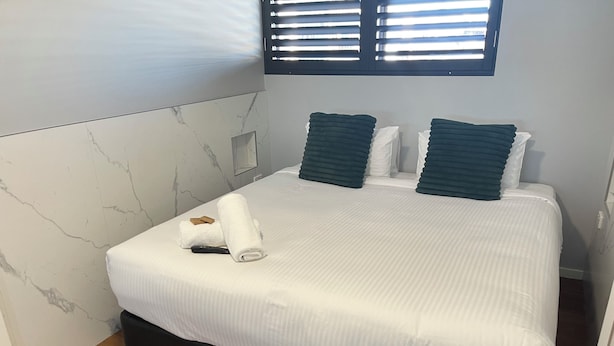 A neatly made bed with white bedding and two dark pillows, a wooden floor, and a marble-patterned wall.