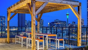 Una estructura de pérgola de madera con bancos en una terraza en la azotea con vistas a un paisaje urbano al atardecer.