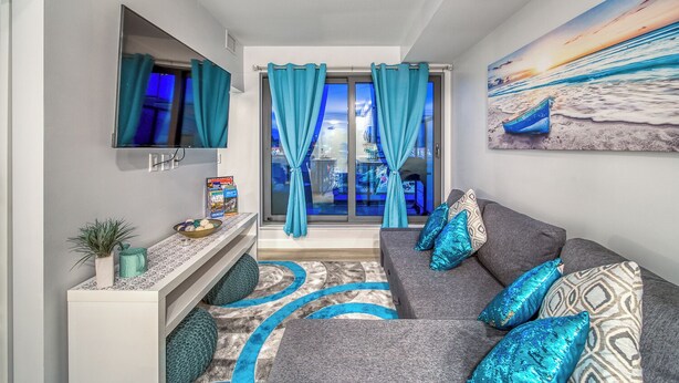 Un salón moderno con un sofá gris, cojines color turquesa, un televisor de pantalla plana y un cuadro con una playa.