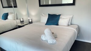 A hotel room with two beds, white linens, a teal pillow, a white towel, and a mirror above the beds. - Standard Two Bedroom (Newcastle)