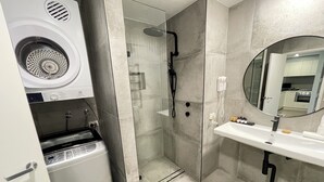 A modern bathroom with a built-in washing machine, a glass-enclosed shower, and a vanity with a mirror. - Prestige Two Bedroom (Newcastle)