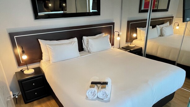 A hotel room with a large bed, two bedside tables with lamps, a mirror above the bed, and a wooden floor. - Deluxe One Bedroom Apartment (Newcastle)