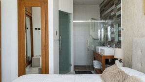 Een moderne slaapkamer met een bed, nachtkastje, spiegel en een badkamer met een douchecabine van glas en een wastafel.