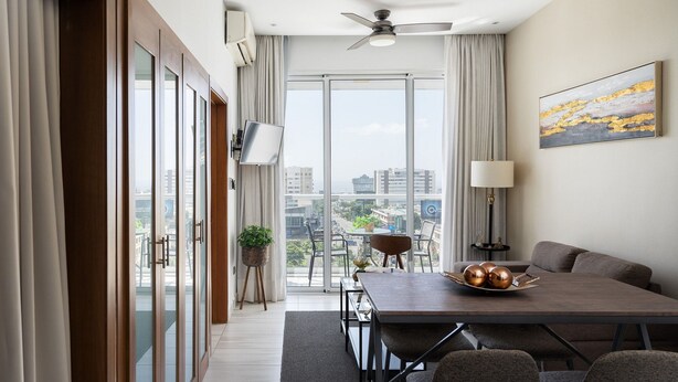 Een moderne hotelkamer met een eethoek, grote ramen, een plafondventilator en uitzicht op de stad.