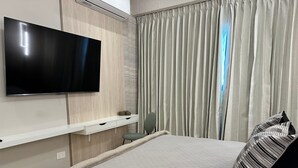 Ein modernes Hotelzimmer mit Wandfernseher, Nachttisch mit Lampe, einem Bett mit gestreifter Bettwäsche und einem großen Fenster mit Vorhängen.
