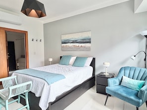 Quarto com cama, mesas de cabeceira, uma cadeira, escrivaninha e um quadro na parede.
