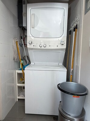 A compact laundry room with a front-loading washing machine, a drying rack, and a trash bin.