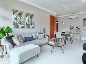 A modern living room with a sofa, a dining table, and chairs. There is a large plant and artwork on the wall.
