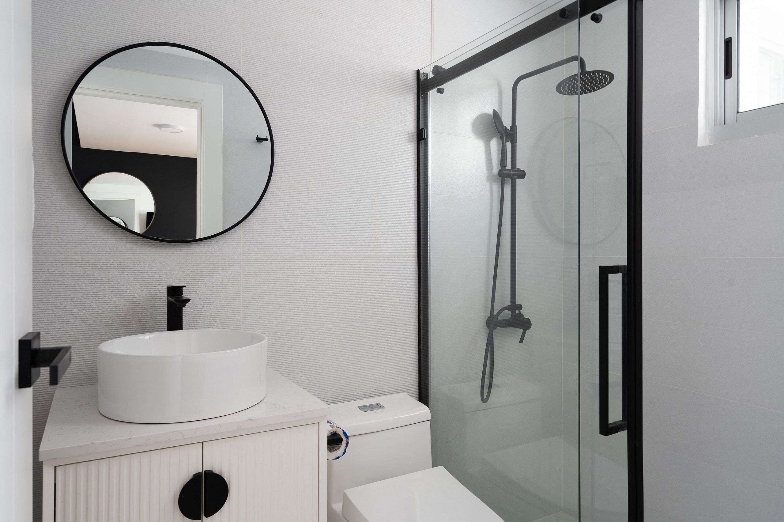 A modern bathroom with a round mirror, a white sink, a glass-enclosed shower, and a toilet.