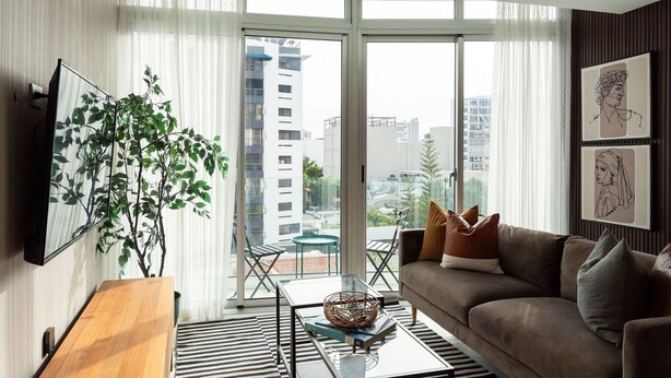 A modern living room with a sofa, coffee table, and a large window with a view of buildings. - 1BR Piantini King Suite Near Blue Mall, Pool & Sauna | YellowKey (santo domingo)