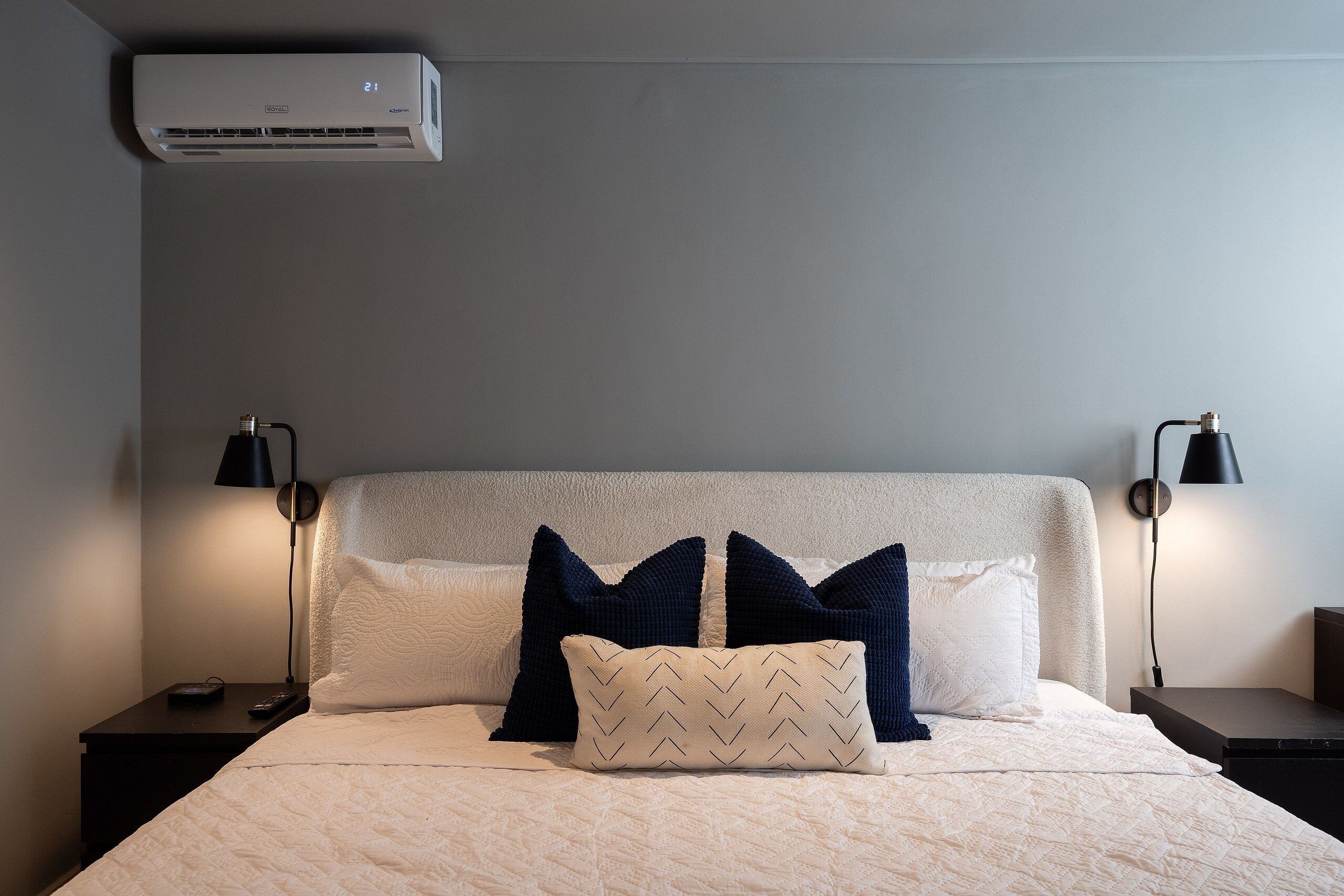 A neatly made bed with a white quilt, two navy blue pillows, and a patterned pillow. There are two bedside tables with lamps, and an air conditioning unit above the bed.