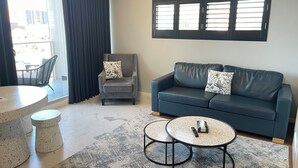 Un salon moderne avec un canapé gris, deux tables rondes, un fauteuil et une grande fenêtre équipée de stores.