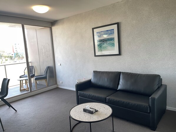 A modern living room with a black leather sofa, a small round table with a tissue box, a framed picture on the wall, and a sliding glass door leading to an outdoor area with chairs. - Standard One Bedroom (Newcastle)