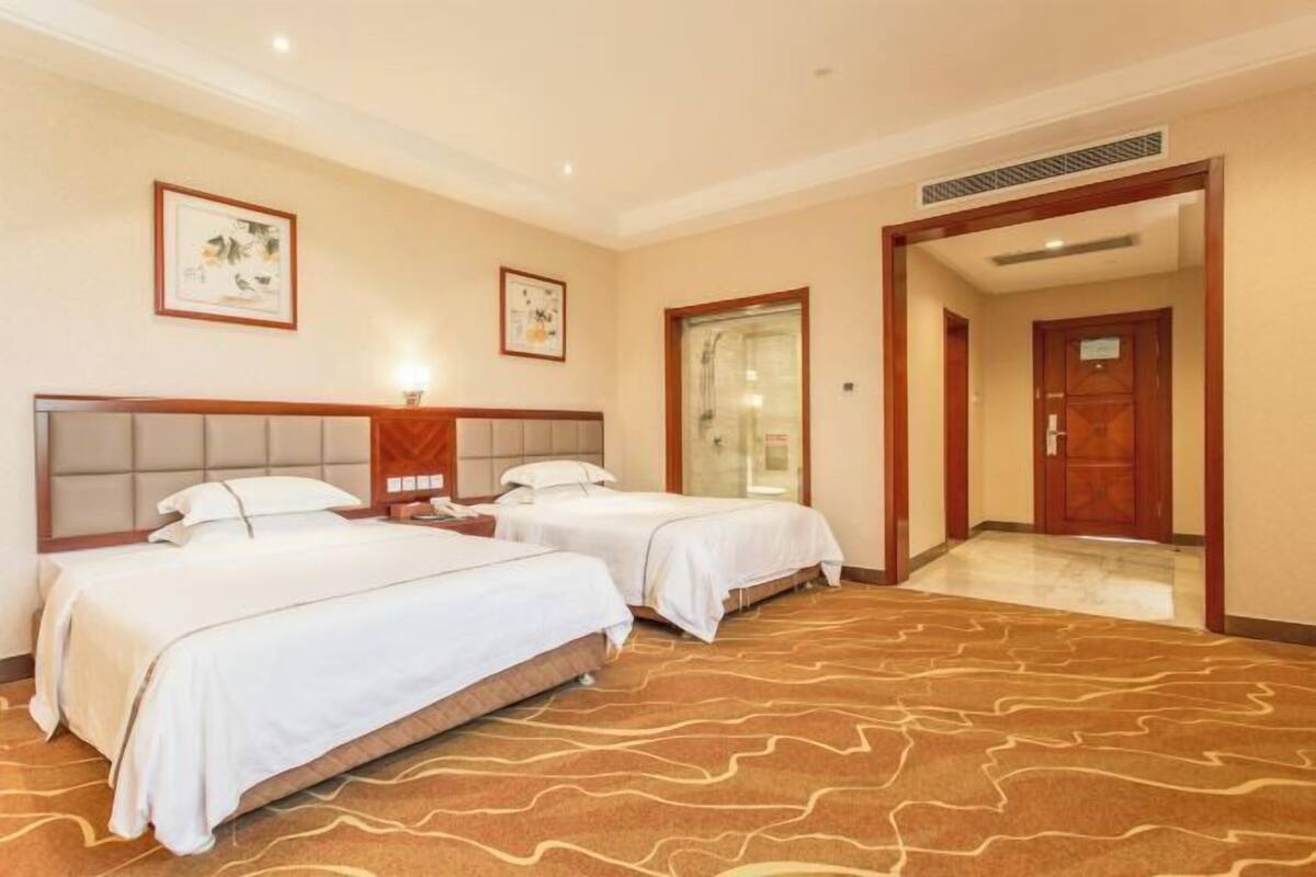 A hotel room with two beds, a wooden headboard, a patterned carpet, and framed artwork on the walls.