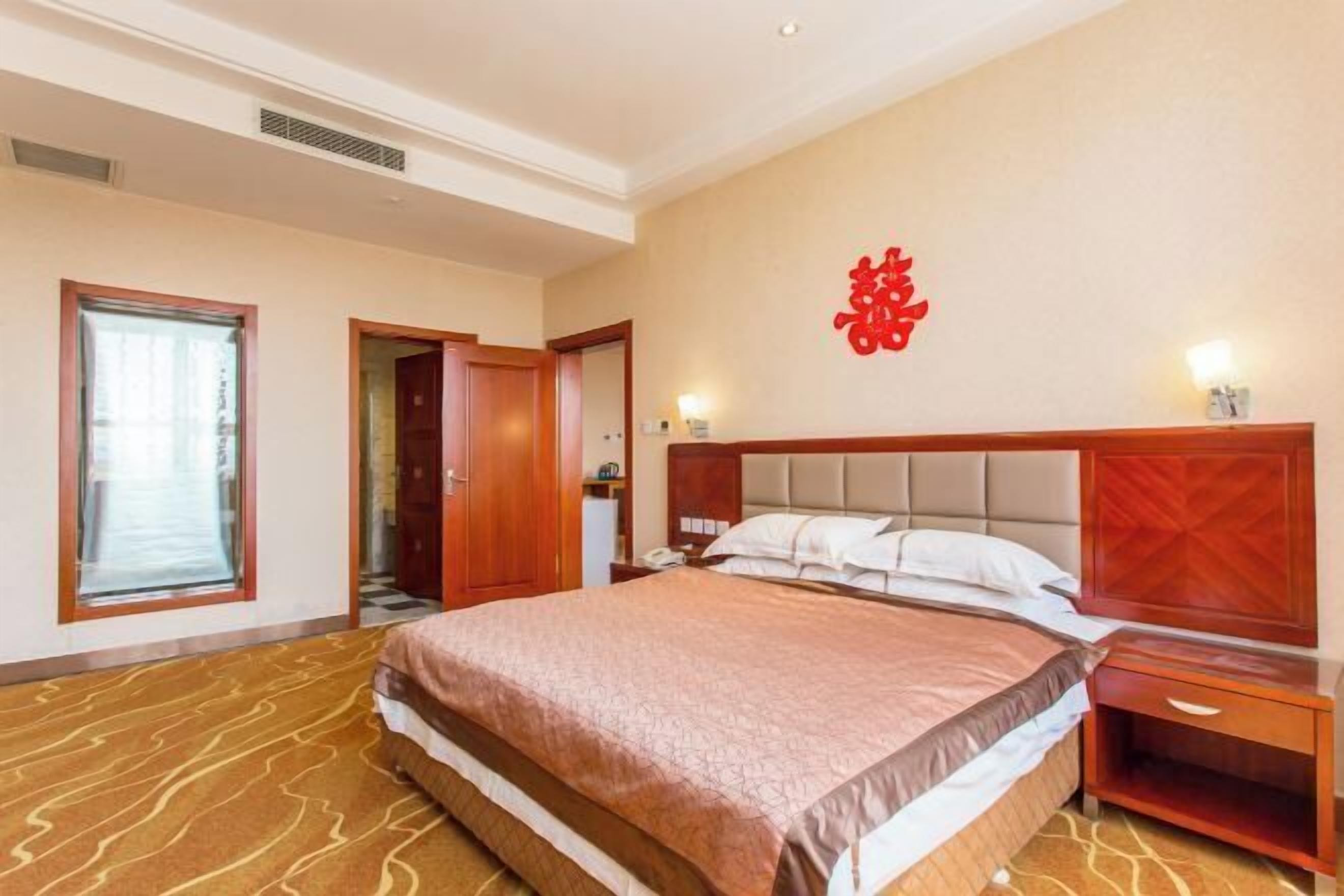 A hotel room with a large bed, a wooden headboard, a nightstand, and a wall with a red decorative element.