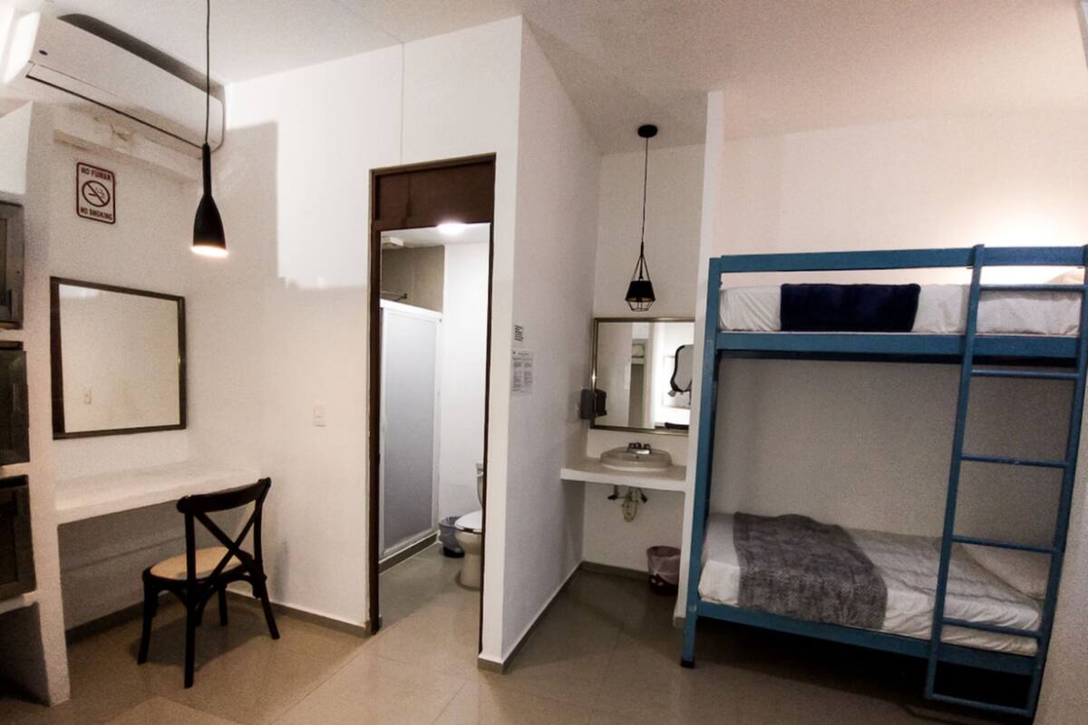 A compact room with a bunk bed, a desk with a chair, a small bathroom, and a hanging light fixture.