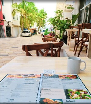 Un menú de restaurante con opciones de comida y precios, una taza blanca, y sillas y mesas de madera.