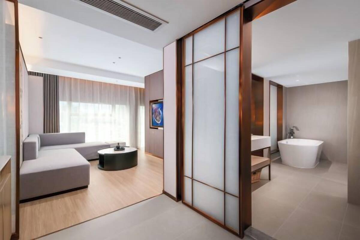 A modern hotel room with a sofa, a round coffee table, a large window with curtains, a flat-screen TV, a bathtub, and a sliding door.