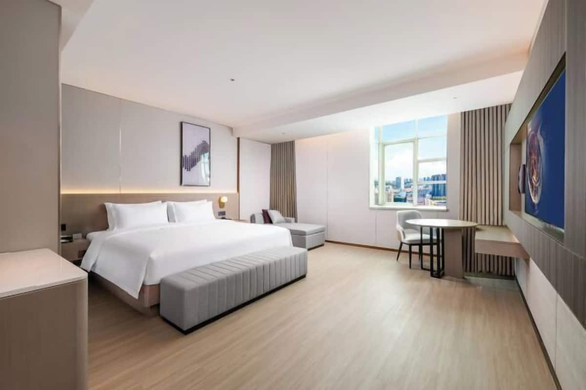 A hotel room with a large bed, a desk, a chair, a window with a city view, and a mounted artwork.
