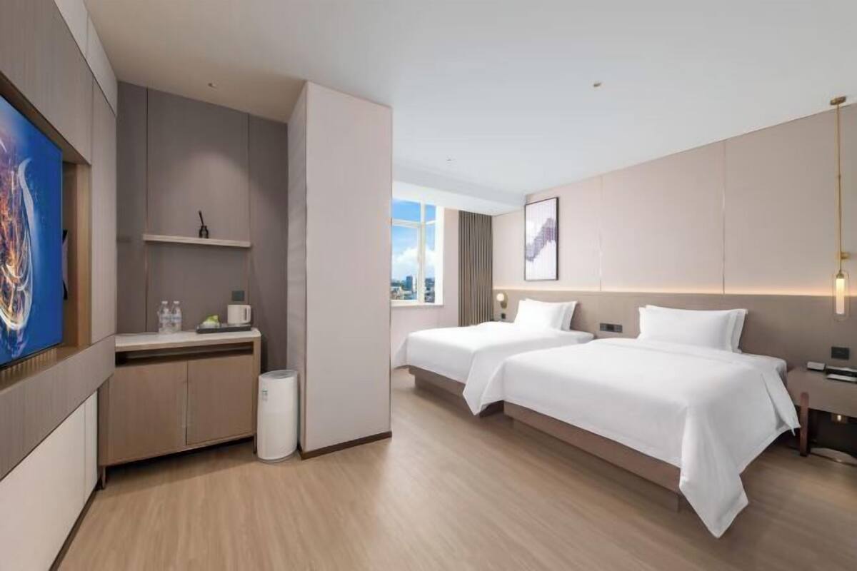 A modern hotel room with two beds, a desk, a small kitchenette, and a view of the city.