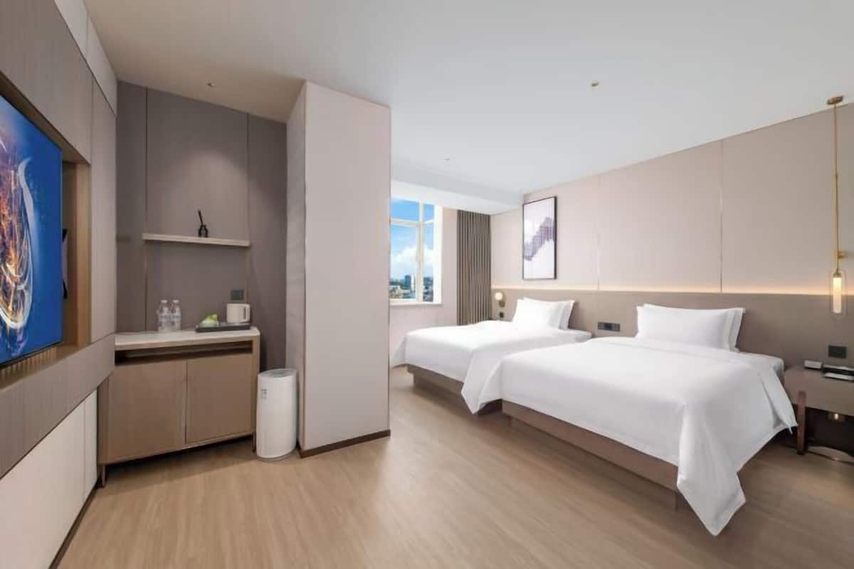 A modern hotel room with two beds, a desk, a small kitchenette, and a view of the city.