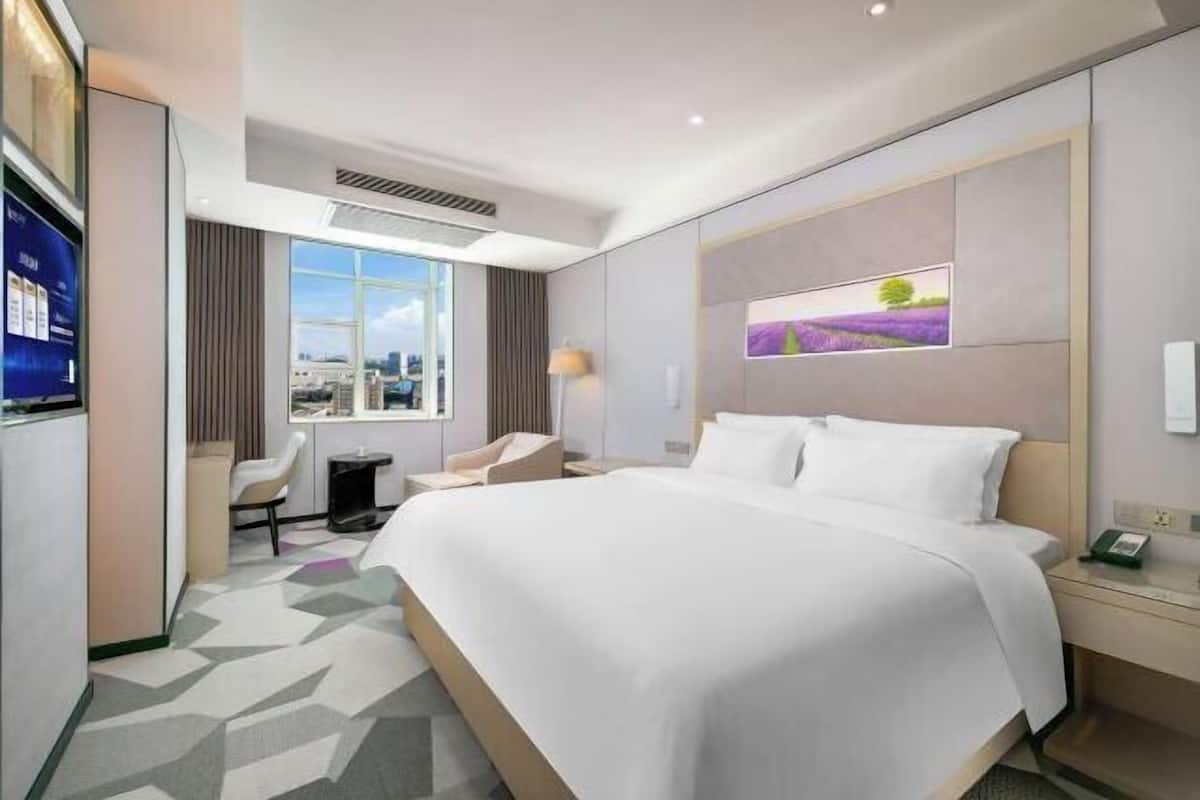 A hotel room with a large bed, a desk, a chair, a window with a city view, and a painting on the wall.