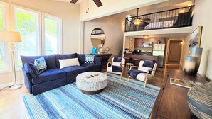 A living room with a dark blue sofa, a round coffee table, and a patterned rug. There is a ceiling fan, a dining area with bar stools, and a kitchen with a microwave and cabinets. - Better to WAKE at the LAKE (Hot Springs)