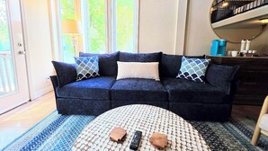 A dark blue sofa with patterned pillows, a round coffee table with remote controls, and a view of a balcony with a railing. - Better to WAKE at the LAKE (Hot Springs)