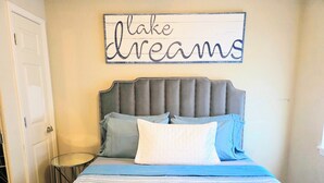 A bedroom with a bed, pillows, a bedside table, and a wall sign. - Better to WAKE at the LAKE (Hot Springs)