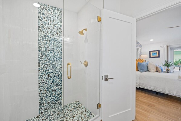 Un baño moderno con pared de ducha de mosaico, cabina de ducha con paredes de vidrio y una puerta blanca que da a un dormitorio con cama y ventana.