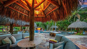 A tropical outdoor lounge area with thatched roof seating, a pool, and lush greenery. - Kon-Tiki Falls: Jupiter`s Exclusive Private Resort! (Jupiter)