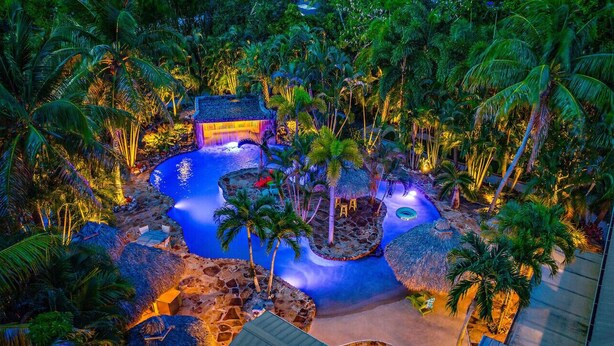 A tropical resort with a central pool surrounded by palm trees and thatched-roof structures. - Kon-Tiki Falls: Jupiter`s Exclusive Private Resort! (Jupiter)