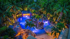 Un resort tropical con una piscina central rodeada de palmeras y estructuras con techo de paja.