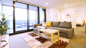 A modern living room with a sofa, a coffee table, and a large window offering a view of the city. - 2bd 2bth - Chic Apt in Woden CBD (Phillip)