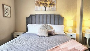 Un lit bien fait, recouvert d’une couette grise, d’un plaid rose et agrémenté de coussins décoratifs. Un tableau représentant un arbre est accroché au mur, au-dessus du lit.