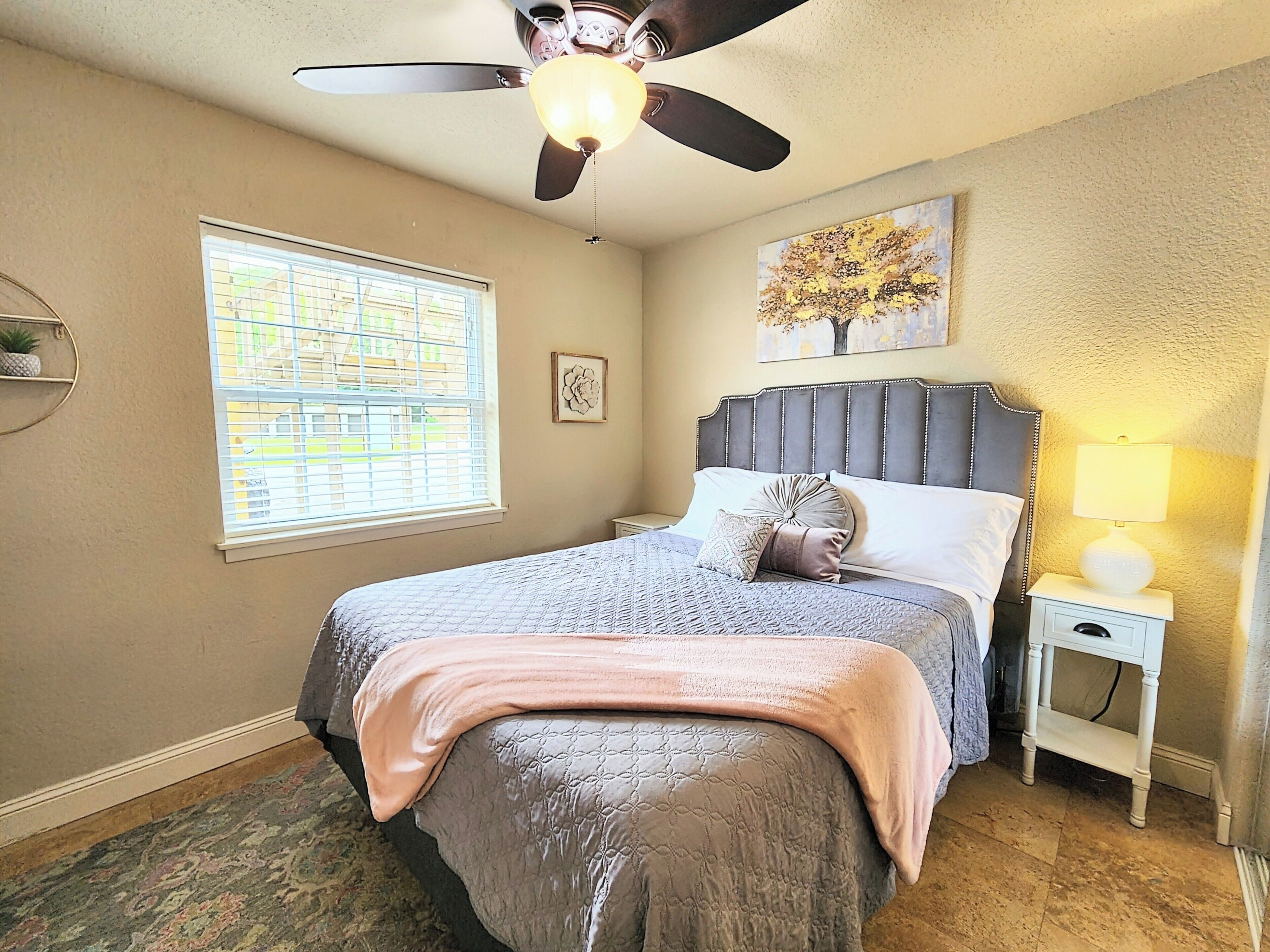 A bedroom with a bed, a ceiling fan, a window with blinds, a nightstand with a lamp, and a painting on the wall.