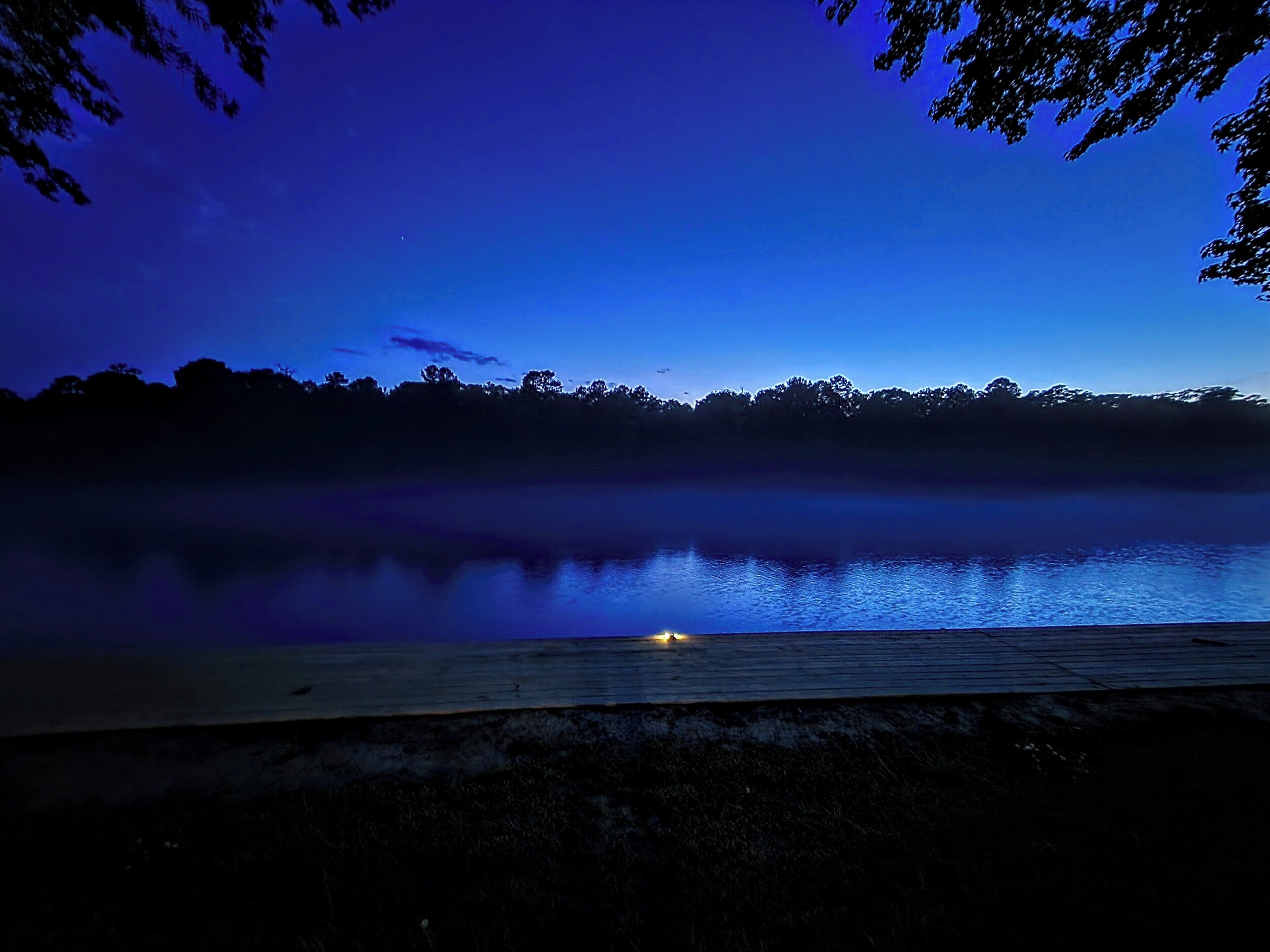 A nighttime view of a calm lake with a misty horizon and a distant tree line.