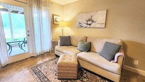 Un salon avec un canapé beige, des coussins à motifs, un pouf en bois, un tapis, une lampe et un tableau accroché au mur.
