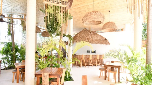An indoor dining area with wooden tables and chairs, surrounded by tropical plants and thatched ceiling lamps. - Muuk Tulum by The Spot Rentals (Tulum)
