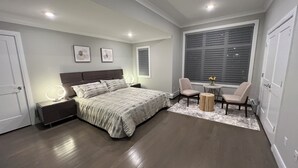 A modern bedroom with a large bed, bedside tables, a vanity with a mirror, two chairs, a small round table with flowers, and a large window with blinds.