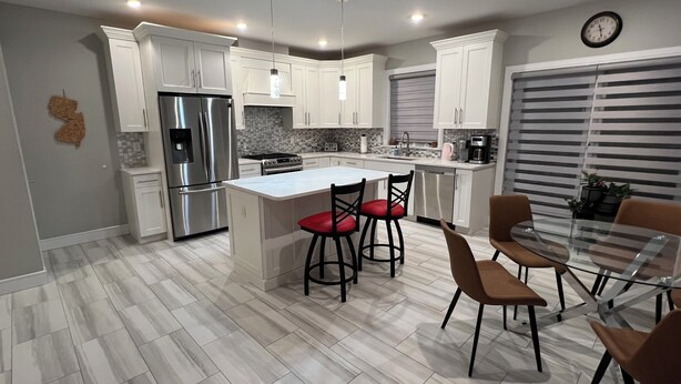 A modern kitchen with white cabinetry, a stainless steel refrigerator, a central island with red bar stools, and a dining area with a glass table and brown chairs.