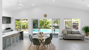 Eine moderne Küche mit Essbereich, Blick auf einen Pool und einem Wohnzimmer mit Sofa und Pflanze.